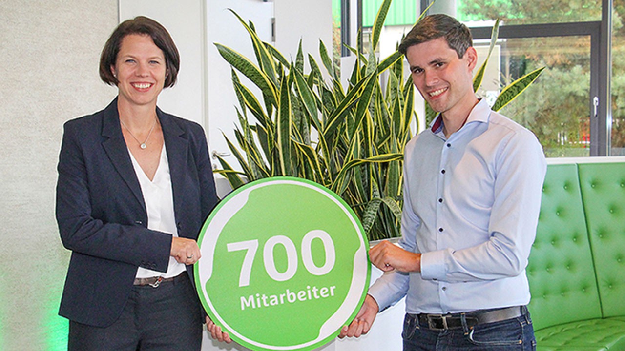 Eine Frau und ein Mann halten ein grünes rundes Schild mit der Aufschrift "700 Mitarbeiter"